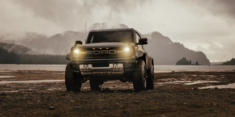 A 2025 Ford Bronco Sport with headlights on, parked along a beach shoreline with forest trees and mountains in the background under a cloudy sky.