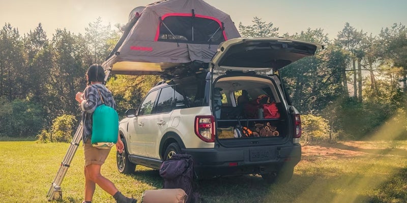 A man walking away from a tan 2024 Ford Bronco Sport equipped with a roof rack with a tent secured and other camping gear visible inside the vehicle